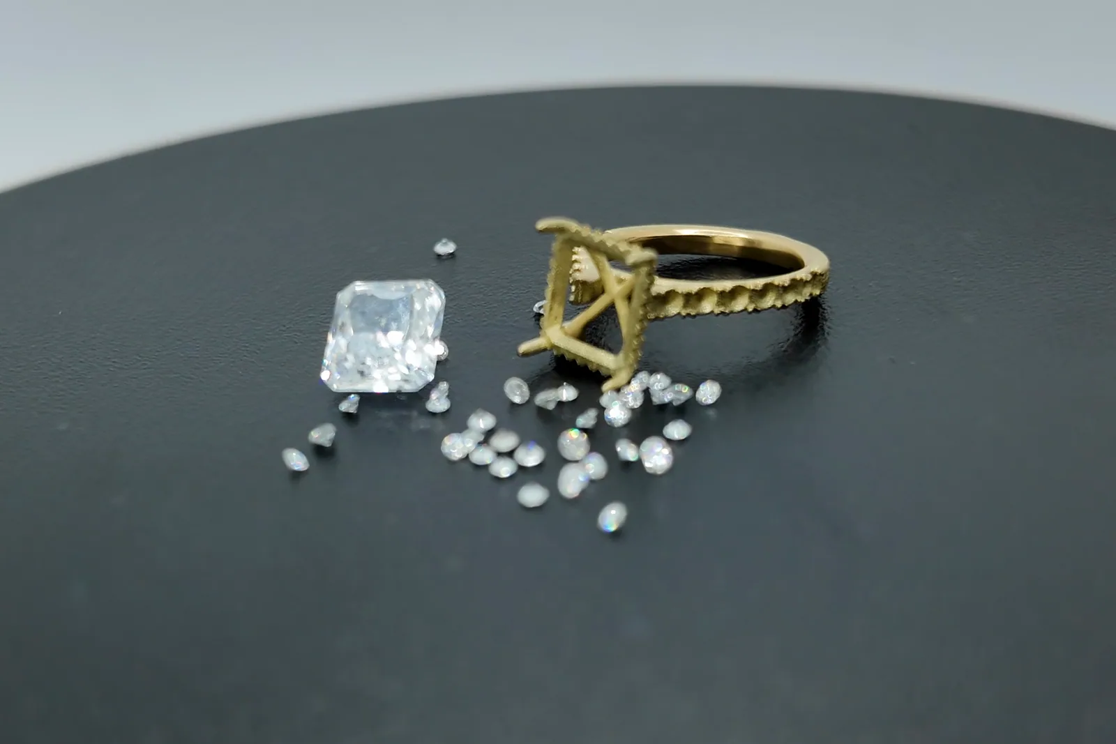A gold ring setting beside loose diamonds and a large emerald-cut stone on a black display stand