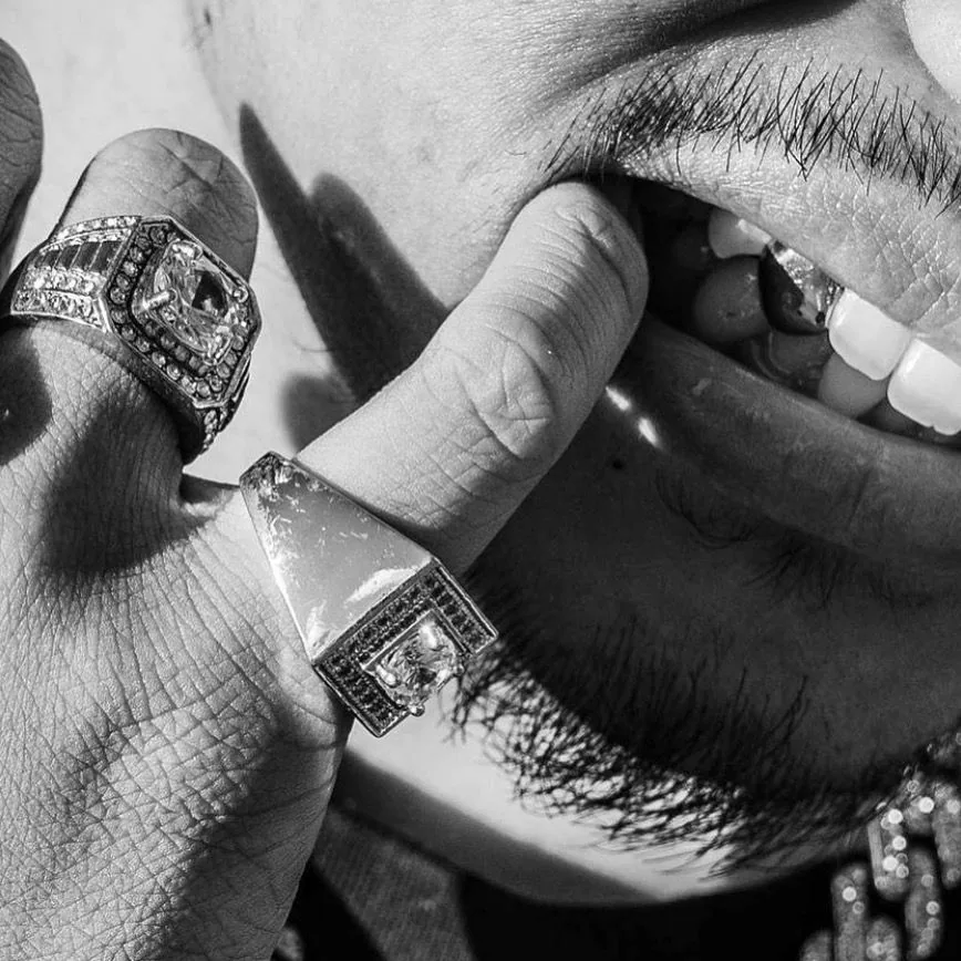 Black and white close-up of a hand with two large stone-set rings held to a smiling mouth, with a grillz tooth visible