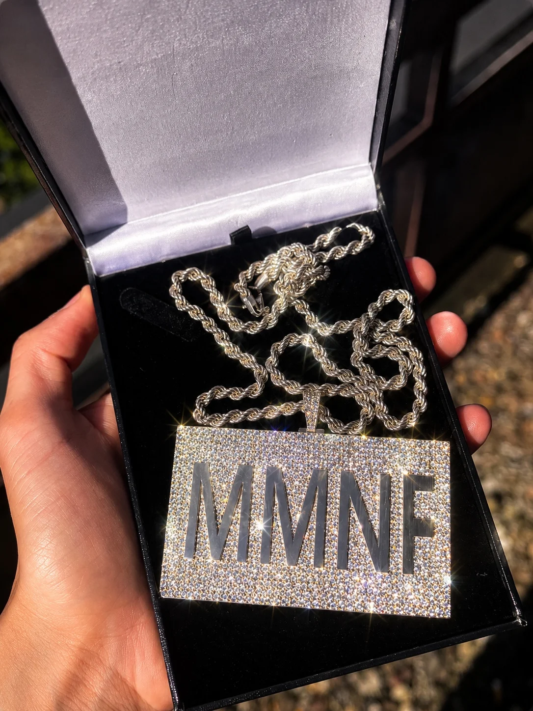A rectangular stone-set pendant with the letters MMNF cut out, on a rope chain in an open black presentation box