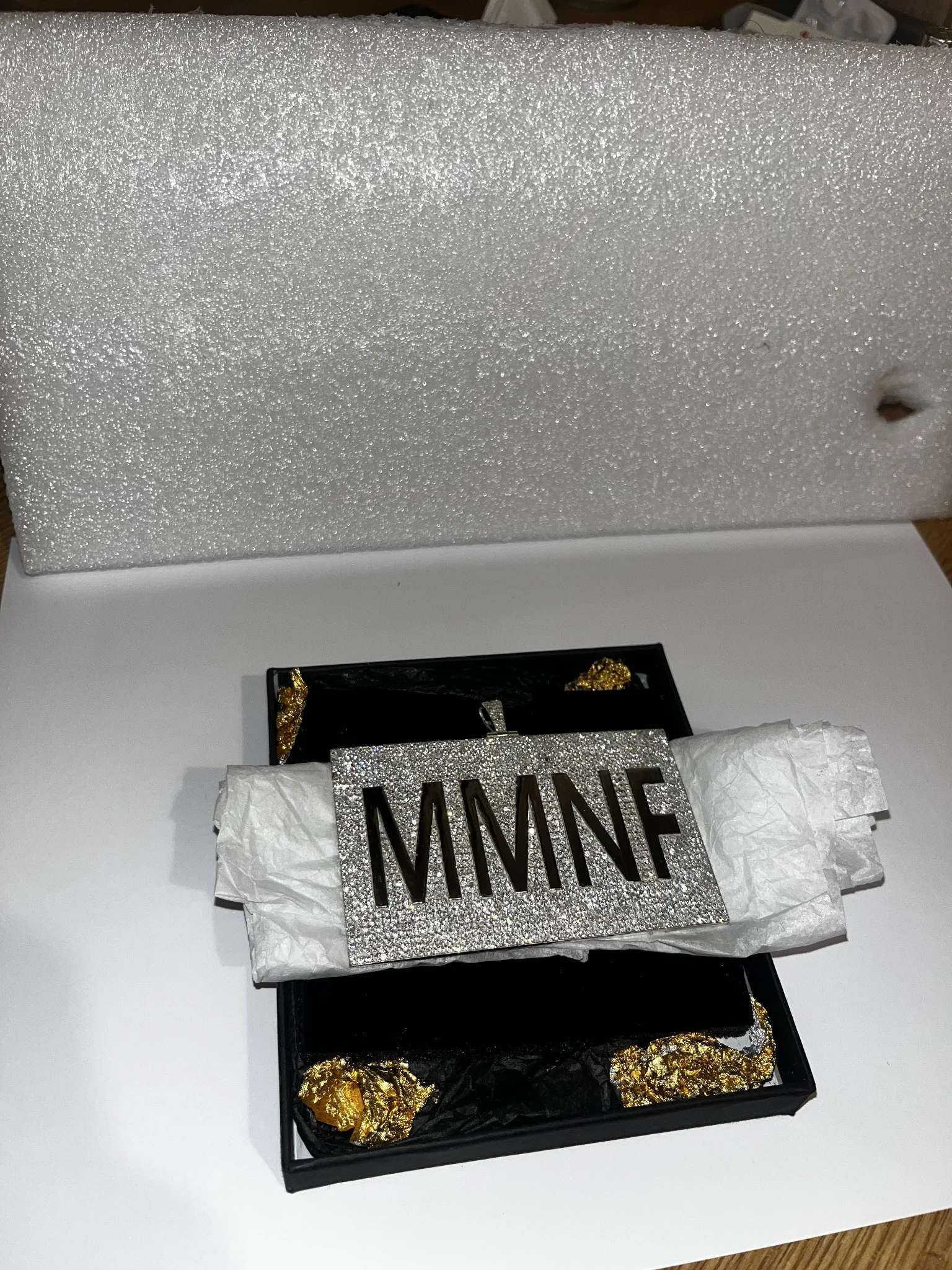 A rectangular stone-set pendant with the letters MMNF cut out, resting on tissue paper in a black presentation tray