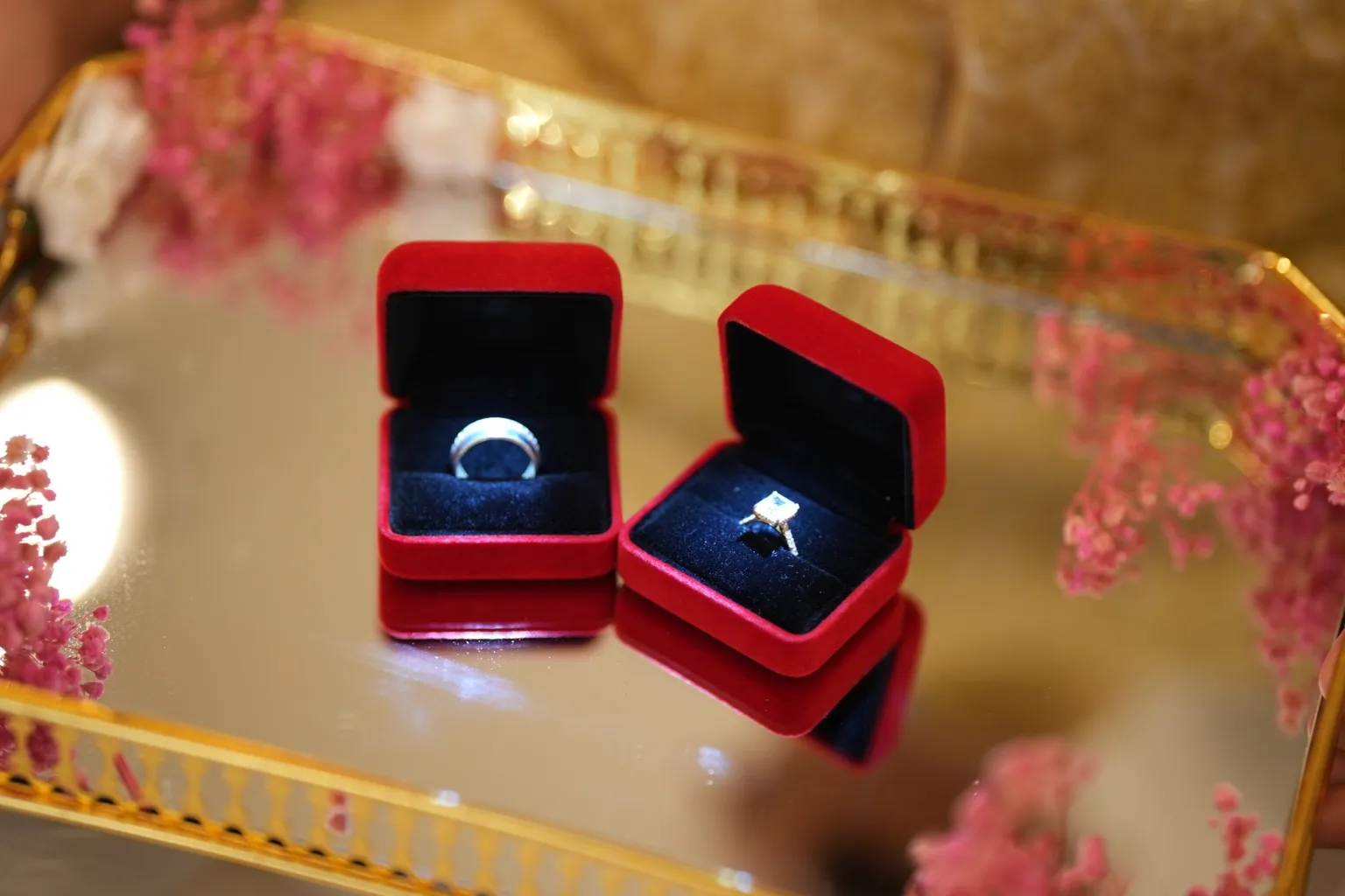 An engagement-style ring and a plain band, each in its own open red velvet presentation box, on a mirrored gold tray with pink flowers