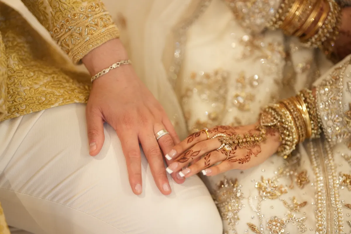 Two hands resting side by side at a wedding, one wearing a silver band and a chain bracelet beside a hand decorated with henna, rings and gold bangles